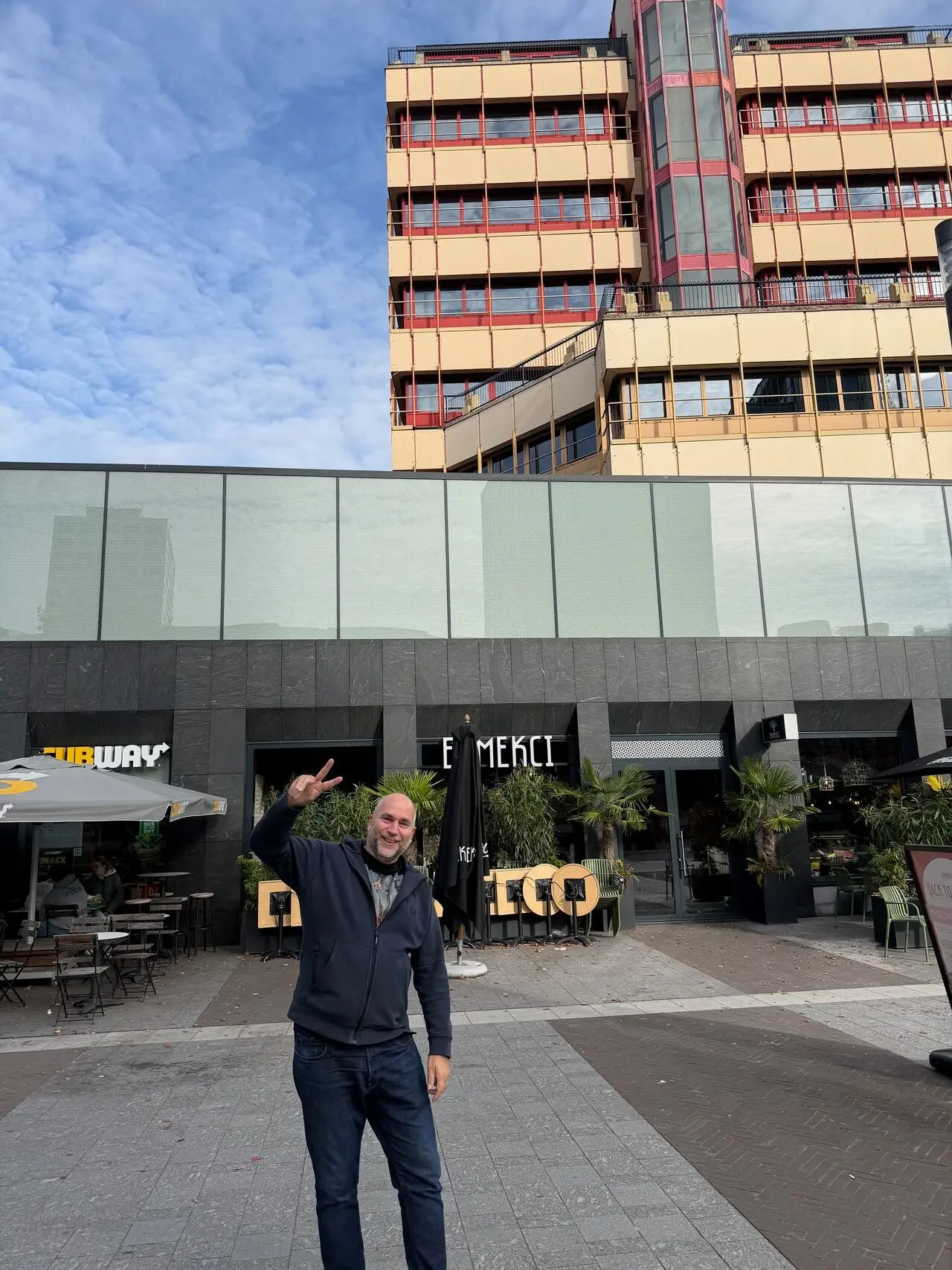 Man standing outside modern building with glass facade and tall structure behind, making peace sign. Building features storefronts with Subway and Merci signs, surrounded by outdoor seating and plants under blue sky.